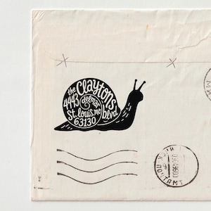 May include: A rectangular off-white envelope featuring a black ink stamp of a snail with the text 'the Claytons 4443 delmar St. louis.mo 63130 blvd' inside the shell. Two circular postmarks and three horizontal wavy lines are printed in black ink.