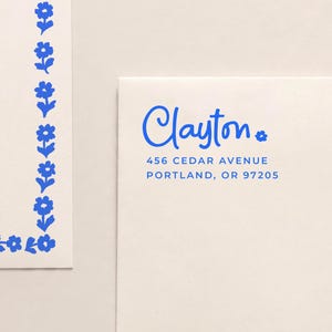 May include: Off-white stationery featuring a blue floral border and a matching envelope printed with the name Clayton, the address 456 Cedar Avenue, Portland, OR 97205, and a small blue flower icon next to the name in a blue sans-serif font.