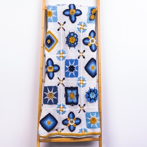 May include: A white crocheted blanket with blue and yellow geometric patterns hanging on a wooden ladder.