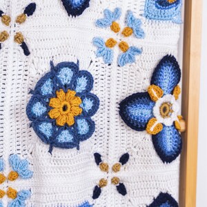 May include: A white crocheted blanket with blue and yellow flowers. The blanket is hanging on a wooden dowel.