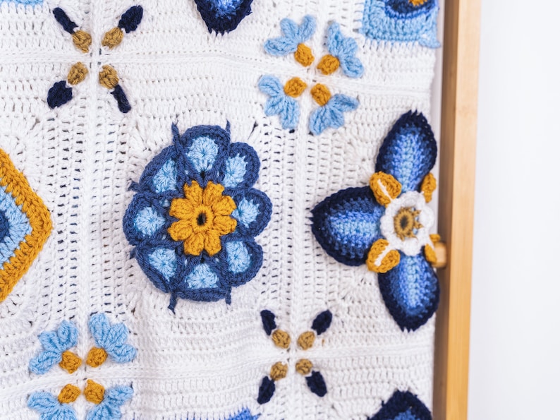 May include: A white crocheted blanket with blue and yellow flowers. The blanket is hanging on a wooden dowel.