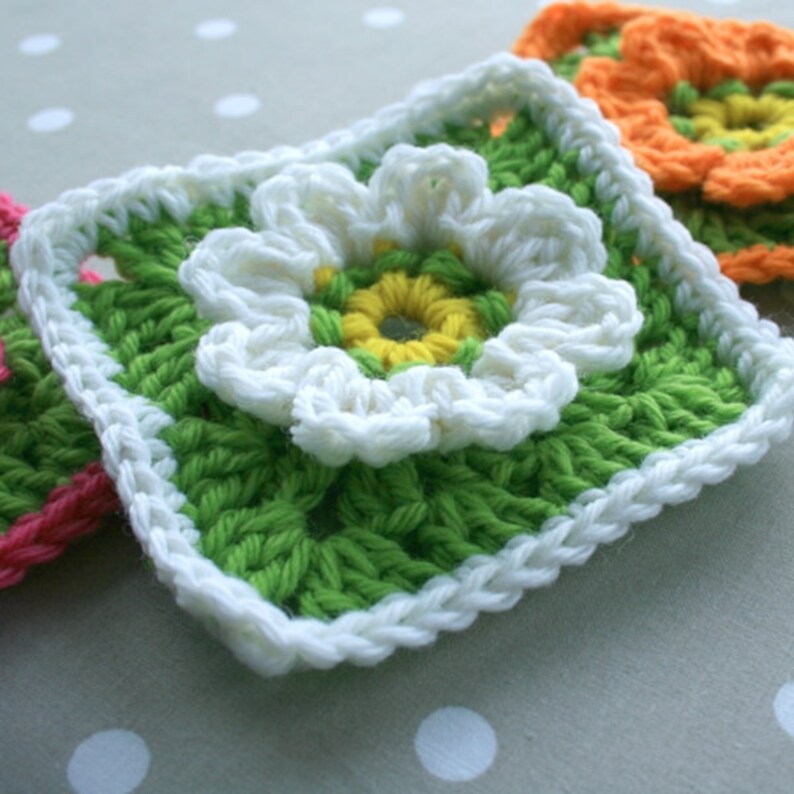 May include: A green crochet square with a white flower in the center. The flower has a yellow center and white petals.