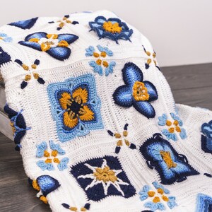 May include: A white crocheted blanket with a blue and yellow floral pattern. The blanket is folded and draped over a wooden crate.