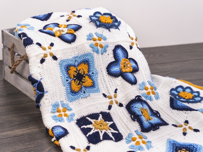May include: A white crocheted blanket with a blue and yellow floral pattern. The blanket is folded and draped over a wooden crate.