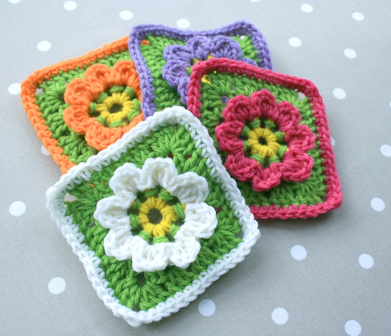 May include: Five crocheted flower squares in different colors. The squares are made with a granny stitch pattern and feature a central flower motif. The colors are orange, pink, purple, green, and white.