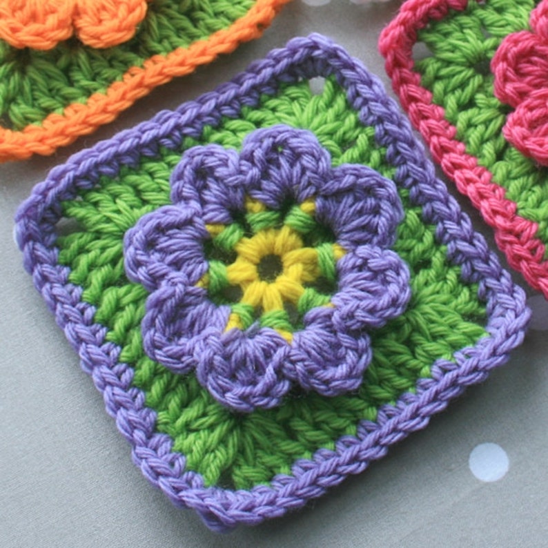 May include: A crocheted flower square with a purple flower in the center, surrounded by green and yellow crocheted layers.