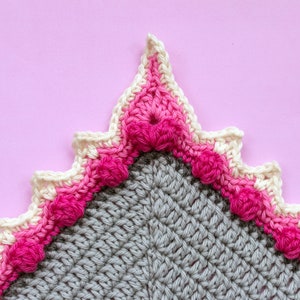 Peut inclure: Un gros plan sur le coin d'une couverture au crochet avec un bord festonné. La couverture est faite de fil gris, blanc et rose. Le bord est décoré d'une rangée de boules roses.