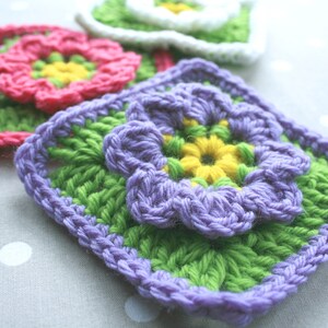 May include: Three crocheted squares with different colored flowers. The first square has a pink flower with a yellow center. The second square has a purple flower with a yellow center. The third square has a white flower with a yellow center.