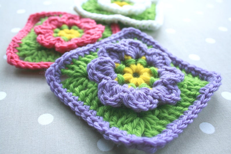 May include: Three crocheted squares with different colored flowers. The first square has a pink flower with a yellow center. The second square has a purple flower with a yellow center. The third square has a white flower with a yellow center.