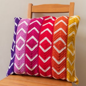 May include: A colorful crocheted pillow with a geometric pattern in shades of purple, pink, red, orange, and yellow. The pillow is sitting on a wooden chair.