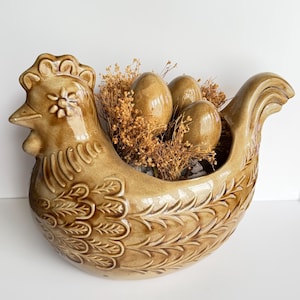 May include: A glossy tan ceramic chicken-shaped planter featuring embossed feather patterns on the body and a flower detail on the head. The interior holds three smooth tan ceramic eggs nestled among dried golden-brown decorative filler plants.