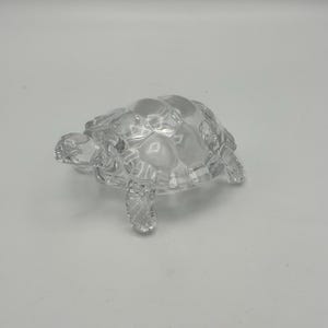 May include: A clear glass figurine shaped like a turtle. The object features a smooth, transparent surface with molded details on the shell representing the scutes, along with four distinct legs and a small head extending from the main body.
