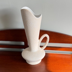 May include: A tall, cream-colored ceramic pitcher vase with a textured, pebbled exterior finish. The vessel features a wide, rounded base, a slender neck that flares upward into a curved, asymmetrical rim, and a single looped handle on the side.