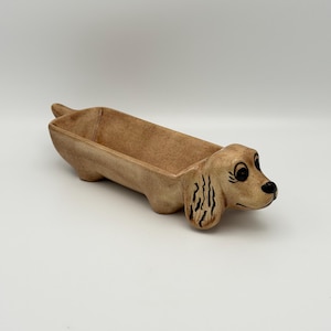 May include: A tan ceramic tray shaped like a long-bodied dog with a textured finish. The dog features painted black eyes, a black nose, and detailed ears. The elongated body forms a hollow, open container suitable for holding small items.