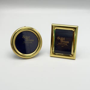 May include: A round and a rectangular photo frame made of polished gold-tone solid brass. Each frame features a dark blue insert with the text Solid Brass PHOTO FRAME (MINI) printed in gold. The frames have a smooth, reflective metallic finish.