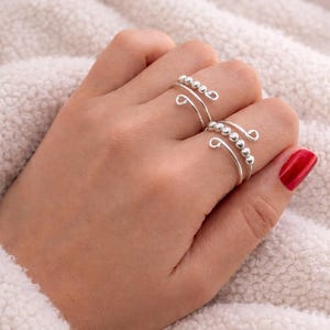 May include: Two silver rings with a spiral design and small beaded accents. The rings are worn on a hand with red nail polish, set against a soft, white background. The jewelry is simple and elegant.