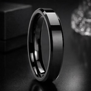 May include: A black ring with a polished, reflective surface and a brushed metal centre band. The ring is set against a dark background, highlighting its sleek design. This piece of jewellery is suitable for everyday wear.