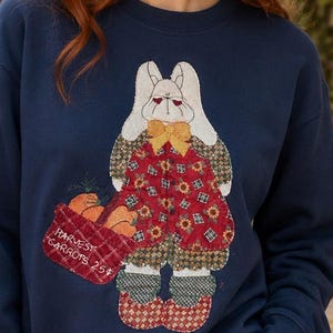 May include: A navy blue long-sleeve sweatshirt featuring a central applique of a white rabbit wearing a red and brown patterned outfit with a yellow bow. The rabbit holds a red basket with the text Harvest Carrots 25 cents printed on it.