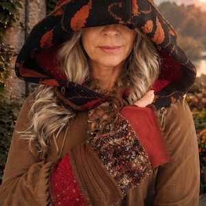 May include: A brown hooded cloak with a patterned hood and matching wrist cuffs. The hood features a black and red design with orange leaf accents. The wrist cuffs have red and brown knitted sections.