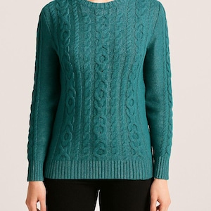 May include: Teal cable-knit sweater with a crew neck. The long-sleeved sweater features a textured cable-knit pattern across the front and sleeves. The model is wearing black pants. The sweater is made of a soft, comfortable material.