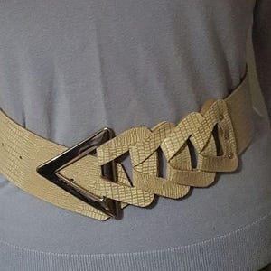 May include: A beige belt with a textured, snakeskin-like pattern. The belt features a silver-toned, triangular buckle and a series of interlocking, triangular decorative elements. The belt is worn around the waist.