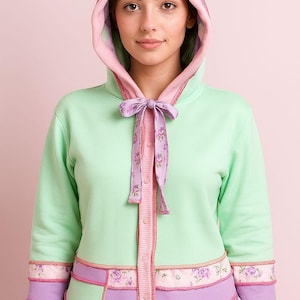 May include: A mint green hooded sweatshirt with a pink-lined hood and floral accents. The sweatshirt features a button-up front with a floral ribbon tie and decorative floral trim at the hem and cuffs. The design incorporates shades of pink and purple.