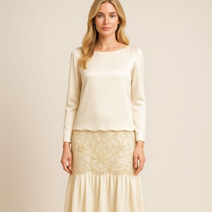 May include: A cream-colored, long-sleeved top with a boat neckline and a matching skirt. The skirt features a detailed, embroidered panel with a scalloped hemline. The outfit is made of a smooth, satin-like fabric, creating an elegant and sophisticated look.