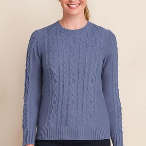 May include: A blue cable-knit sweater with a crew neck. The sweater features a textured pattern on the front and sleeves. The model is wearing dark blue pants. The sweater is a classic style.