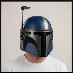 May include: A Mandalorian helmet in shades of blue and gray, with a silver stripe down the center of the dome. The helmet has a T-shaped visor and a vertical antenna-like structure extending from the top.