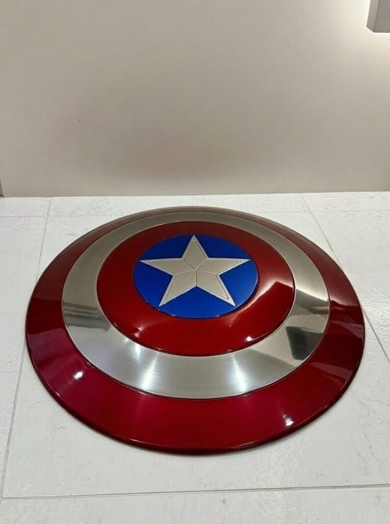 May include: A red, silver, and blue shield with a star emblem. The shield is round with a glossy finish. The outer ring is red, followed by a silver band, and a blue center with a white star.