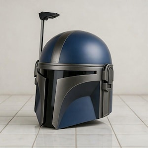 May include: A blue and gray Mandalorian helmet with a black visor and a vertical antenna. The helmet has a rounded top with a gray stripe and angular features. The helmet is displayed on a white tiled surface.