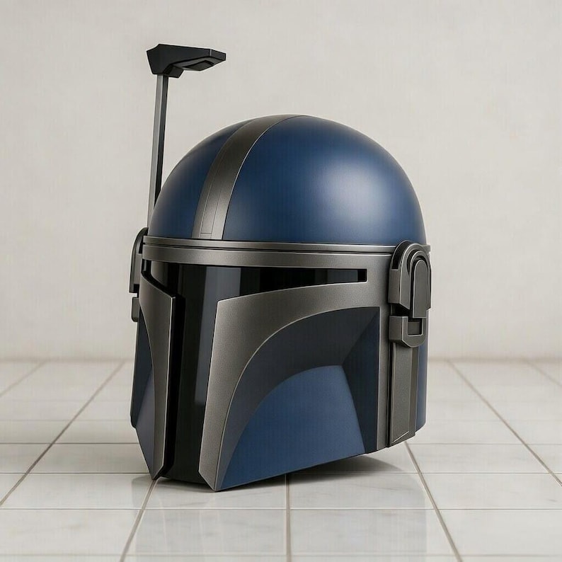 May include: A blue and gray Mandalorian helmet with a black visor and a vertical antenna. The helmet has a rounded top with a gray stripe and angular features. The helmet is displayed on a white tiled surface.