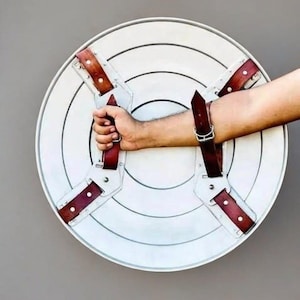 May include: A white, circular shield with concentric circles and brown leather straps. The shield has a handle on the back and is held by a person's arm. The shield is a prop or costume piece.