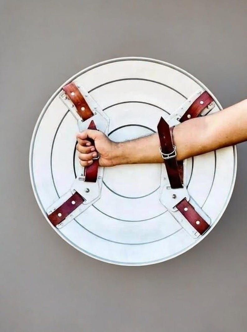 May include: A white, circular shield with concentric circles and brown leather straps. The shield has a handle on the back and is held by a person's arm. The shield is a prop or costume piece.