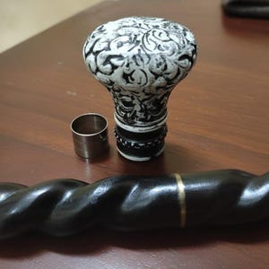 Puede incluir: Un mango de bastón con estampado en blanco y negro con una férula a juego y un eje de madera oscuro y retorcido. El mango tiene forma bulbosa con un diseño detallado. Una banda dorada acentúa el eje.