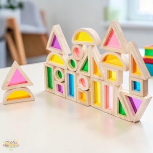 Könnte beinhalten: Holzbausteine in verschiedenen Formen, darunter Dreiecke, Rechtecke und Halbkreise. Die Bausteine haben farbige transparente Einsätze in Pink, Gelb, Grün und Blau. Die Bausteine sind auf einer weißen Oberfläche angeordnet.