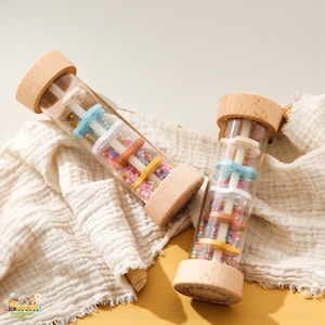 May include: Two clear, cylindrical baby rattles with wooden ends. Each rattle contains colorful beads and pastel-colored discs. The rattles are on a cream-colored cloth, with a yellow background.