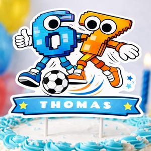 Puede incluir: Una tarta glaseada blanca con glaseado azul y un adorno para tarta con temática de fútbol. El adorno presenta dos figuras de dibujos animados, un balón de fútbol y una pancarta que dice "THOMAS". Dos velas encendidas en el fondo.