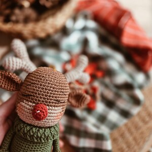 May include: A crocheted reindeer toy featuring a brown head, light beige antlers, and a bright red nose. The reindeer has closed eyes and is wearing a textured olive green sweater with a high collar. The body is crafted from yarn in a dense stitch.