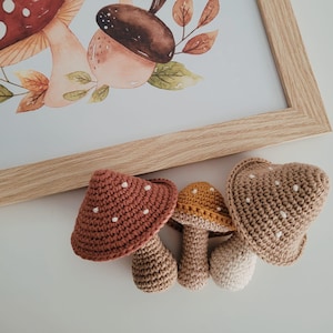 Pode incluir: Uma impressão em aquarela emoldurada com cogumelos e uma bolota, com três brinquedos de cogumelos de crochê em primeiro plano. Os brinquedos são em tons de castanho, bege e amarelo, com bolinhas brancas. A moldura é de madeira clara.