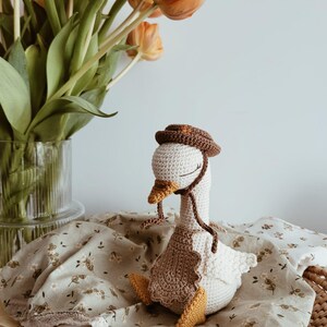 May include: A handcrafted crochet goose toy featuring a cream-colored body, yellow feet and beak, and a brown bonnet tied under the chin. The goose wears a scalloped brown apron over its chest and has embroidered closed eyes with long eyelashes.