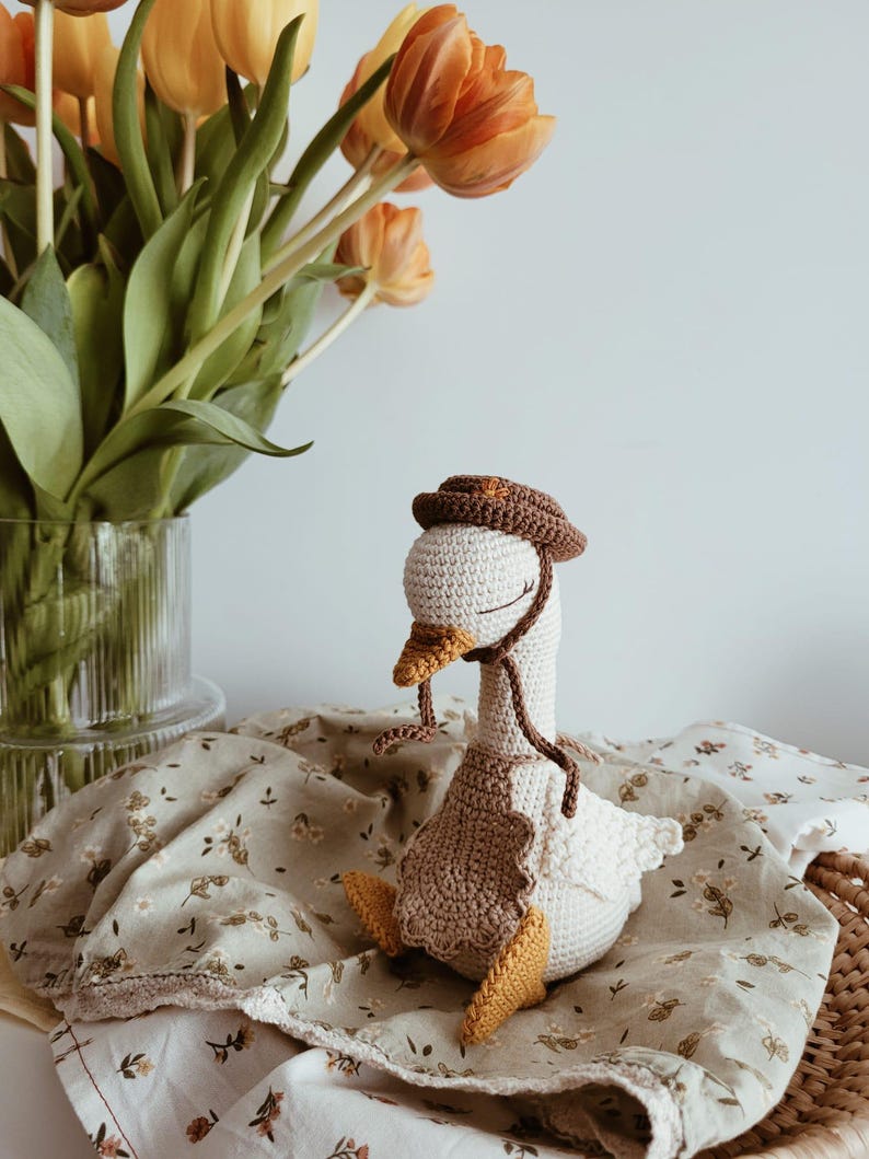 May include: A handcrafted crochet goose toy featuring a cream-colored body, yellow feet and beak, and a brown bonnet tied under the chin. The goose wears a scalloped brown apron over its chest and has embroidered closed eyes with long eyelashes.