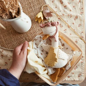May include: A crocheted goose toy with a white body, a yellow beak, and orange feet. The goose wears a pink crocheted bonnet with a floral accent and a yellow crocheted cape. It is nestled inside a square wooden basket lined with floral fabric.