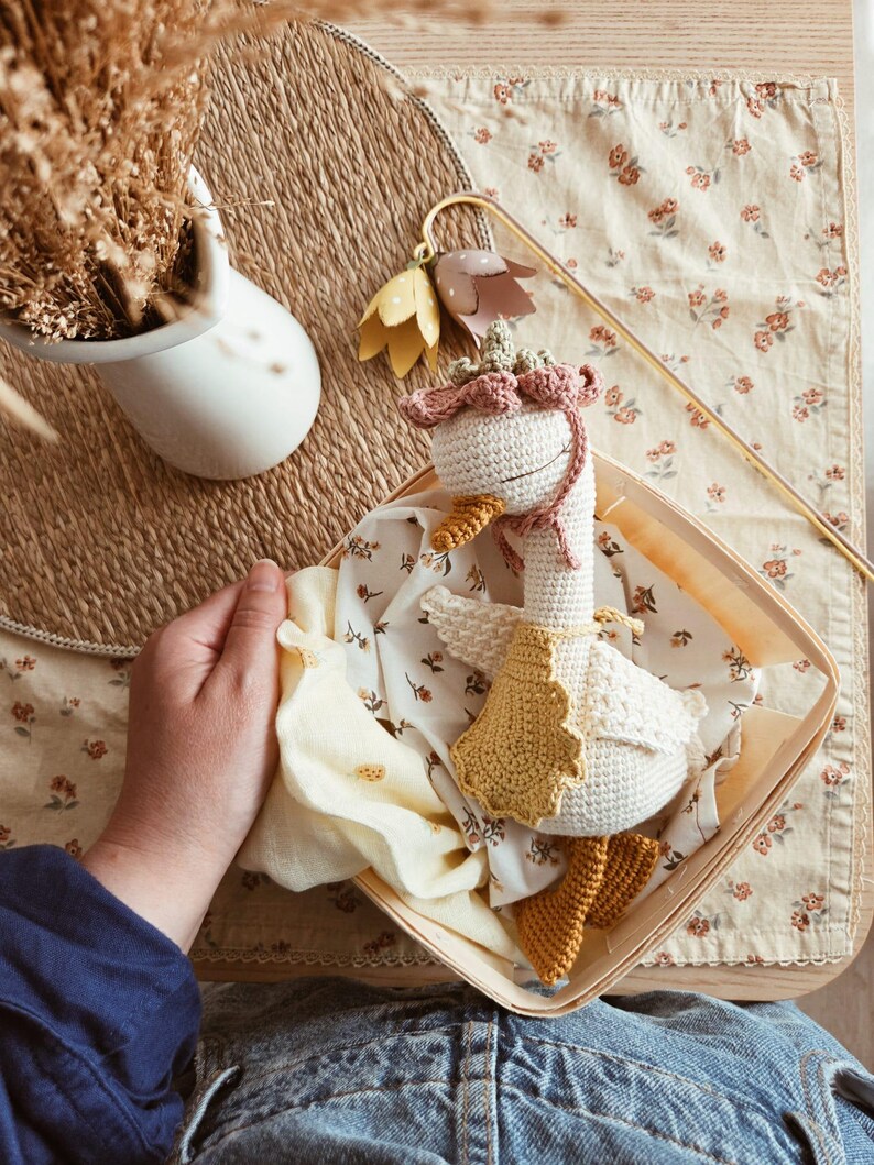 May include: A crocheted goose toy with a white body, a yellow beak, and orange feet. The goose wears a pink crocheted bonnet with a floral accent and a yellow crocheted cape. It is nestled inside a square wooden basket lined with floral fabric.