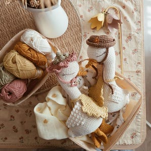 May include: Two handmade crochet geese dolls with closed eyes, wearing small hats and yellow aprons. One goose wears a dusty pink ruffled bonnet, while the other wears a brown hat. They are nestled together in a light-colored wooden basket.