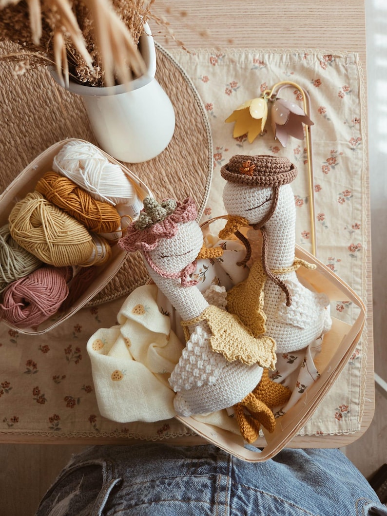 May include: Two handmade crochet geese dolls with closed eyes, wearing small hats and yellow aprons. One goose wears a dusty pink ruffled bonnet, while the other wears a brown hat. They are nestled together in a light-colored wooden basket.