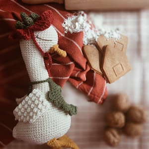 May include: A crocheted goose toy with a cream-colored body, yellow feet and beak, a dark green scarf, and a red and green bonnet. The goose has closed eyes and is positioned near a wooden rolling pin, gingerbread cookies, and walnuts.