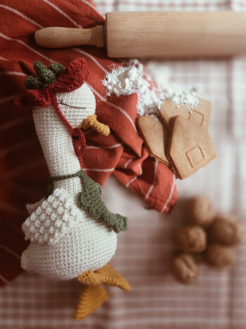 May include: A crocheted goose toy with a cream-colored body, yellow feet and beak, a dark green scarf, and a red and green bonnet. The goose has closed eyes and is positioned near a wooden rolling pin, gingerbread cookies, and walnuts.