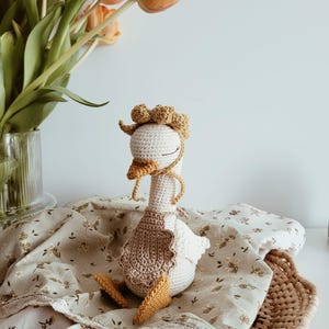 May include: A handcrafted crochet goose toy featuring a cream-colored body, a mustard yellow beak and webbed feet, and a matching mustard yellow bonnet with a ruffled edge. The goose wears a light brown crocheted apron and has closed eyes.
