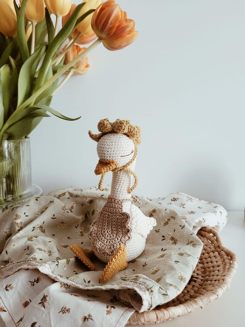 May include: A handcrafted crochet goose toy featuring a cream-colored body, a mustard yellow beak and webbed feet, and a matching mustard yellow bonnet with a ruffled edge. The goose wears a light brown crocheted apron and has closed eyes.